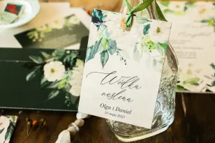 Zawieszki ślubne na butelki weselne na drewnie w kolorze butelkowej zieleni z dodatkiem białych kwiatów i dekoracyjnych gałązek