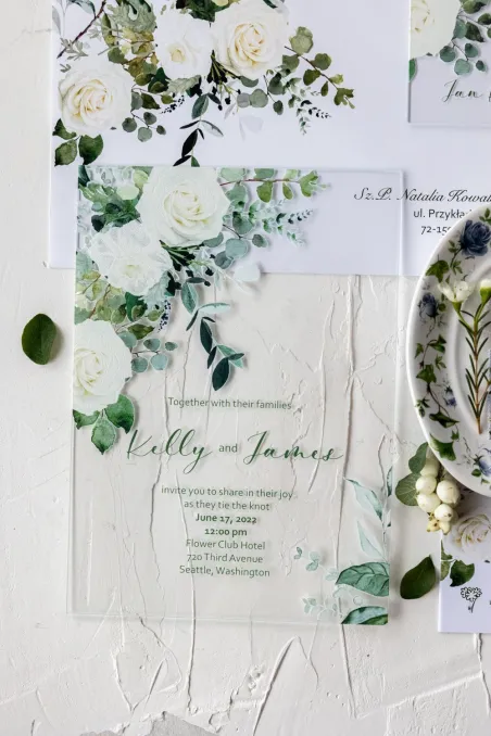 Elegante Hochzeitseinladungen auf Glas mit einem floralen Motiv aus weißen Rosen und grünen Eukalyptuszweigen.