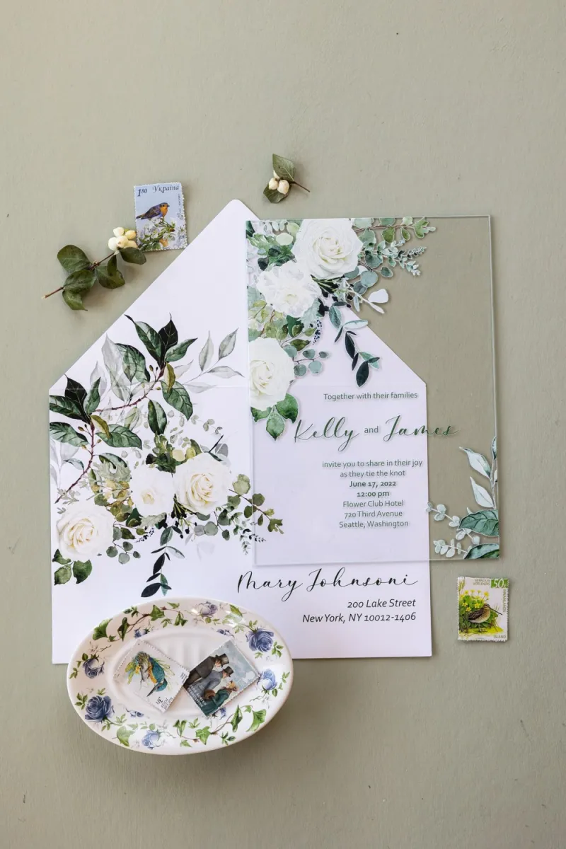 Elegante Hochzeitseinladungen auf Glas mit einem floralen Motiv aus weißen Rosen und grünen Eukalyptuszweigen.