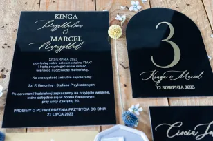 Moderne Hochzeitseinladungen auf Glas mit vergoldeten Vor- und Nachnamen der Braut und des Bräutigams auf schwarzem Hintergrund.