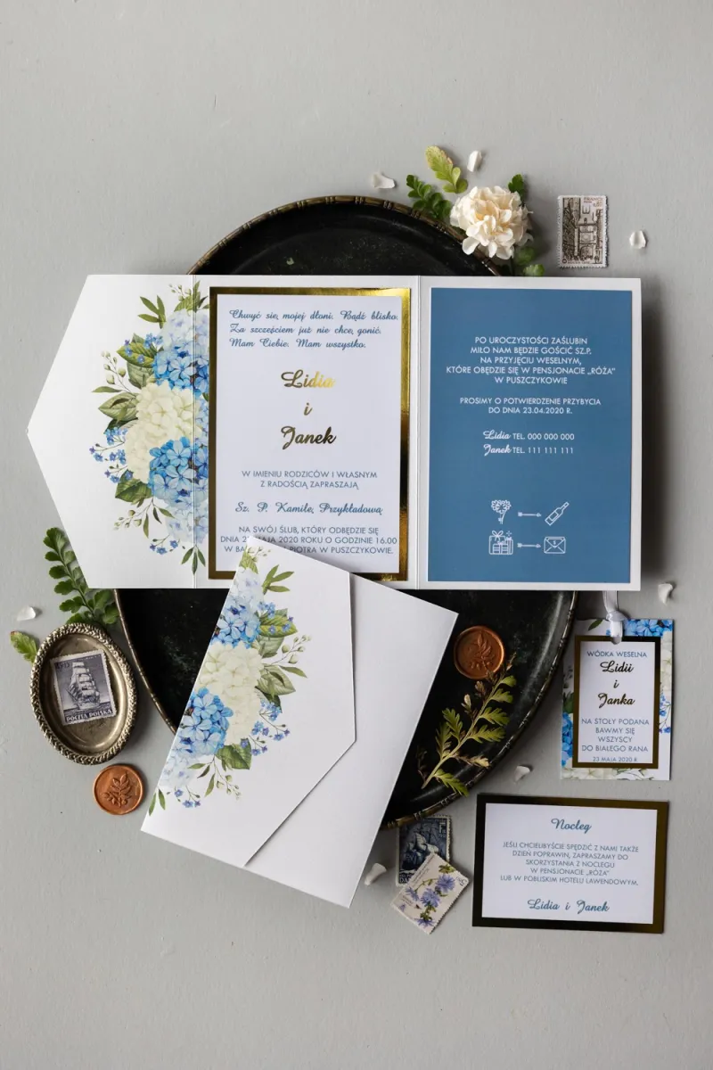 Złote zaproszenia ślubne z niebieską i białą hortensją w otoczeniu zieleni. Zaproszenia ze złotą ramką w eleganckim folderze.