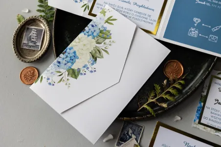Złote zaproszenia ślubne z niebieską i białą hortensją w otoczeniu zieleni. Zaproszenia ze złotą ramką w eleganckim folderze.