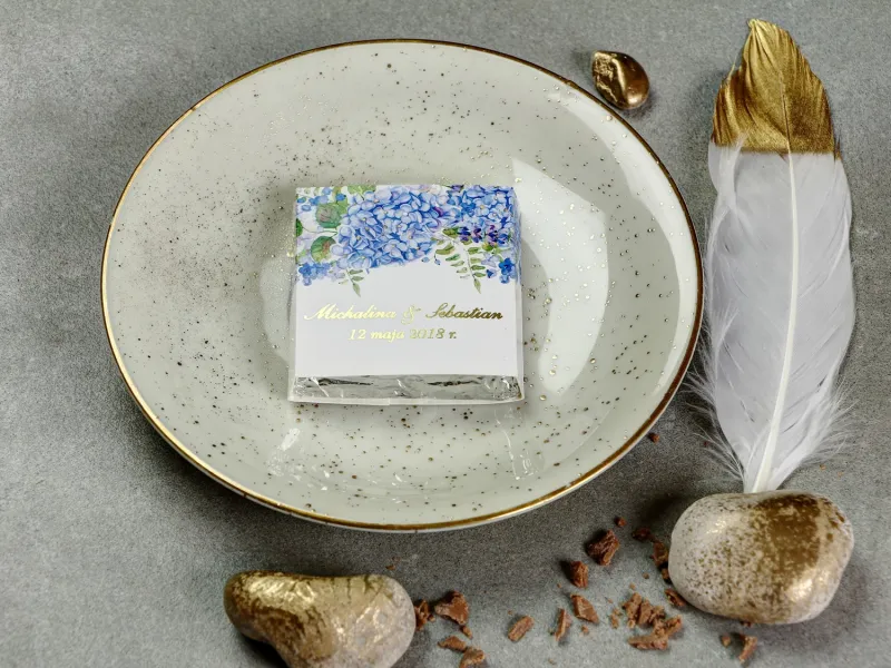 Podziękowanie dla gości weselnych w postaci czekoladki, owijka ze złoconymi napisami oraz grafiką kwiatów hortensji i lawendy