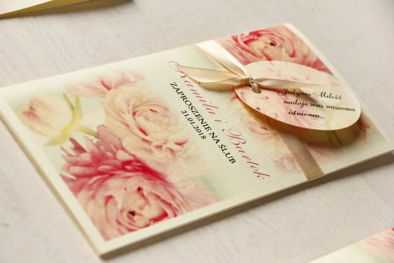 Kremowe zaproszenia ślubne z piwonią w pastelowym odcieniu jasnego różu. Całość przewiązana ozdobną przywieszką z wierszykiem.