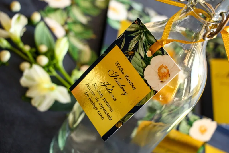Złote zawieszki na butelki weselne w stylu Glamour. Granatowa grafika z białą kamelią i dodatkiem zielonych liści Złote zawieszki na butelki weselne w stylu Glamour. Granatowa grafika z białą kamelią i dodatkiem zielonych liści