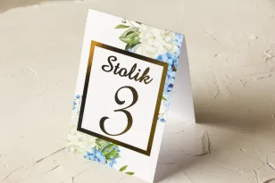 Złocone Numery stolików ślubnych z niebieską i białą hortensją w otoczeniu zieleni