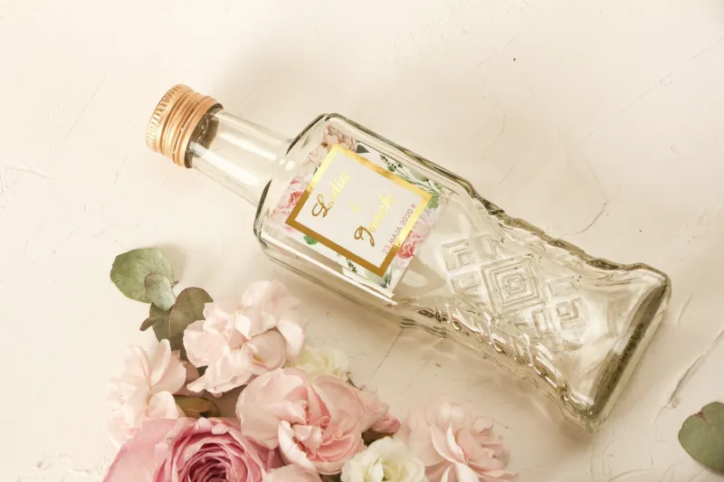 Butelki na nalewki wraz ze złoconą etykietą oraz z różowymi piwoniami w towarzystwie białych róż i zieleni
