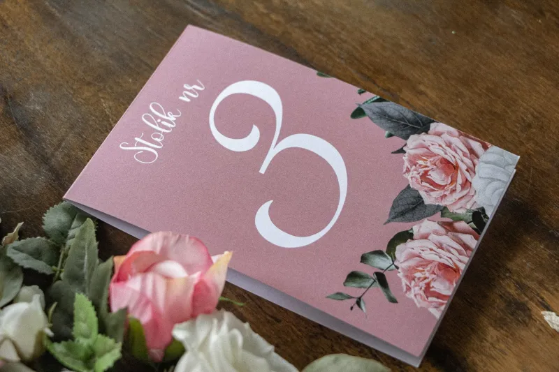 Numery stolików weselnych w kolorze pudrowego różu. Na numerze kompozycja z białych i różowych piwonii i róż