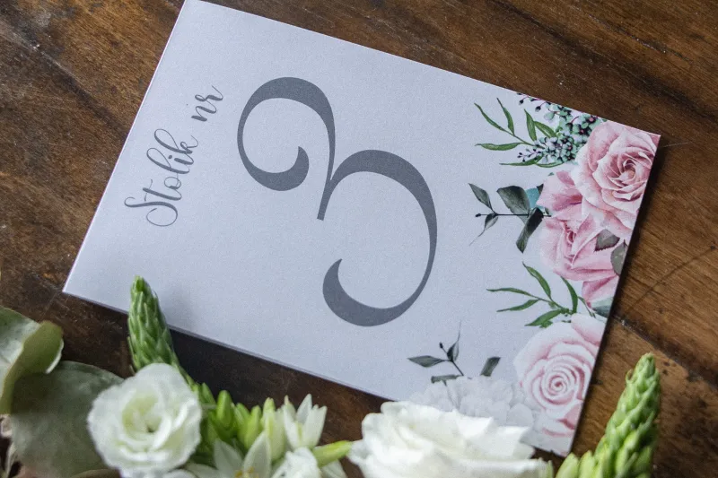 Numery stolików weselnych w kolorze szarym. Na numerze kompozycja z białych i pastelowych róż