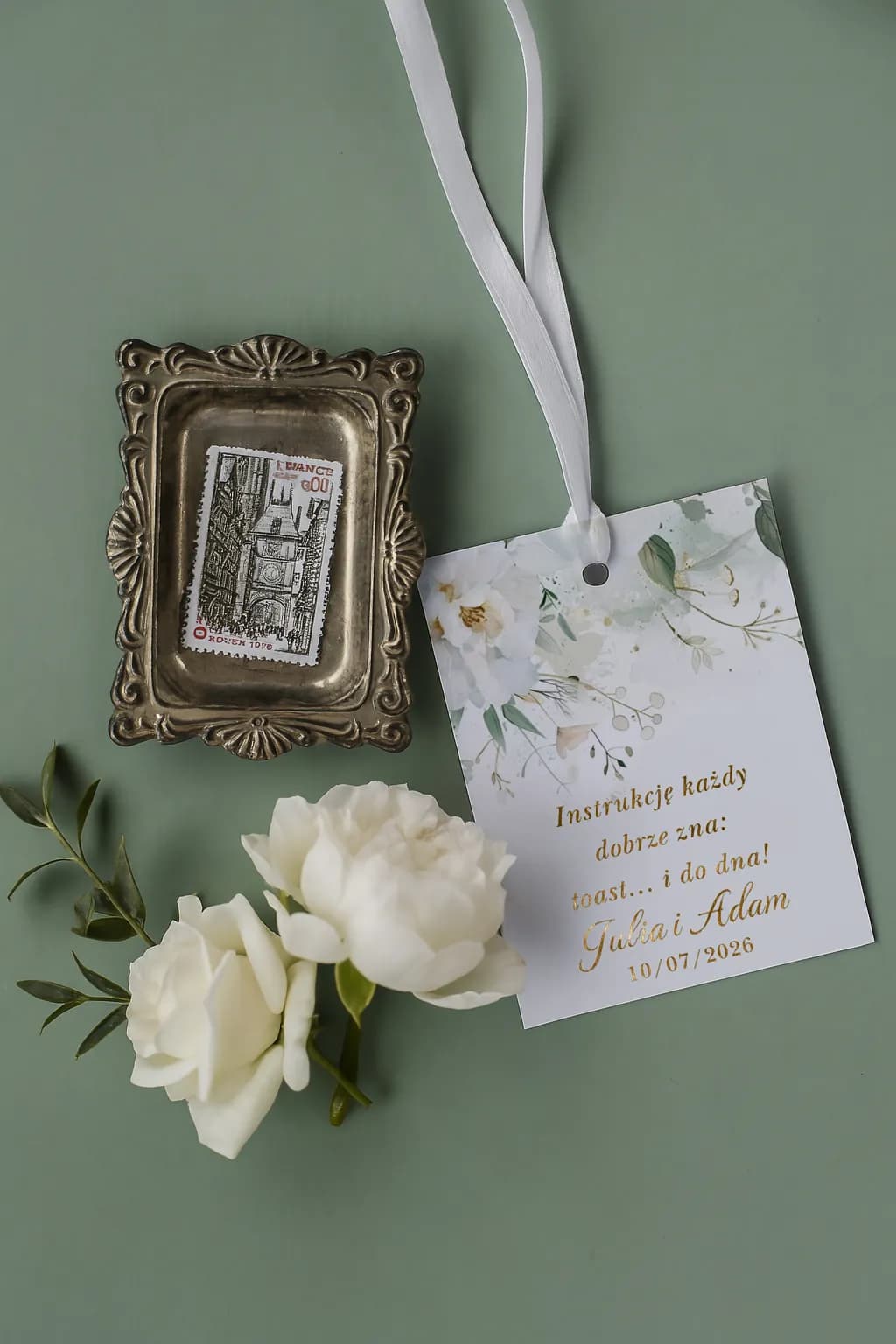 Étiquette de bouteille dorée Ruskus n° 6 Étiquette élégante pour alcool de mariage Ruskus n° 6 avec texte doré, posée sur fond vert à côté de fleurs blanches.