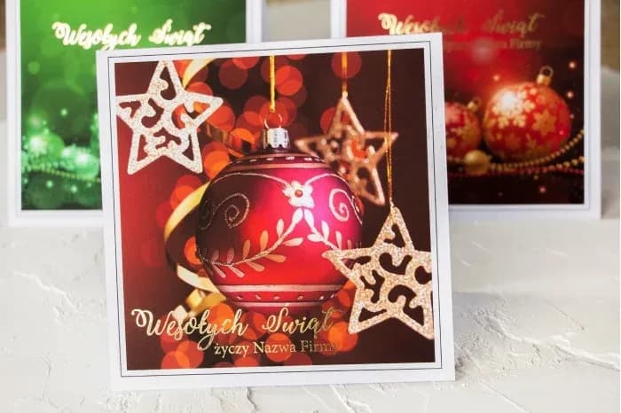 Personalizowana kartka świąteczna z czerwonym motywem bombki i złoconym napisem na satynowym papierze.