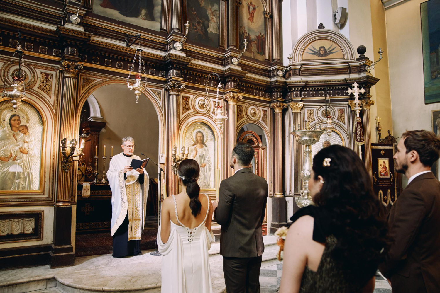 Na zdjęciu drużba i druhna stoją za parą młodą przed księdzem w ikonostasie.